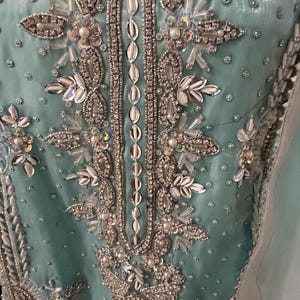 May include: Close-up of a light teal fabric heavily embellished with silver and iridescent beads, sequins, and pearls. The intricate design features floral and geometric patterns, with a central vertical beaded strip. The overall effect is luxurious and ornate.