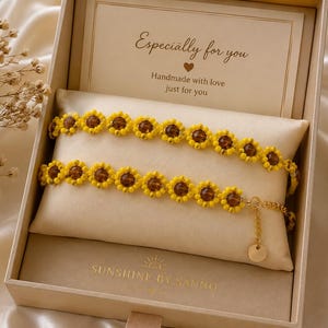 May include: Two beaded bracelets featuring yellow flower-shaped beads with brown centers, presented in a beige gift box. The box includes a card with the text Especially for you and the brand name SUNSHINE BY SANNO printed at the bottom.