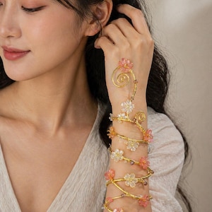 May include: A gold-toned arm cuff with a floral design. The cuff wraps around the arm and features delicate pink and white flowers. The cuff is adorned with small, clear beads and is a decorative accessory.