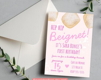 May include: A pink birthday invitation with the text "HIP HIP Beignet!" and "IT'S SARA BINCE'S FIRST BIRTHDAY!". The invitation includes the date, time, location, and RSVP information. A white envelope and greenery are in the background.