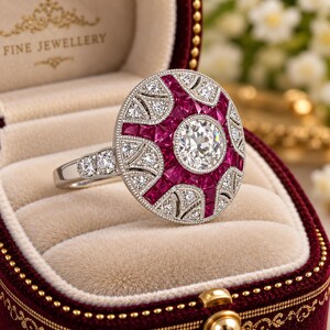 Puede incluir: Un anillo de tono plateado con una gran piedra central redonda y transparente, rodeada por una disposición en forma de estrella de rubíes rojos oscuros de corte cuadrado y piedras transparentes más pequeñas en la banda.