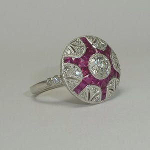 Può includere: Un anello d'argento con una grande faccia rotonda. La faccia è decorata con un diamante centrale circondato da rubini quadrati e diamanti più piccoli. Il gambo presenta piccoli accenti di diamanti.