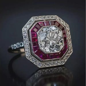 Puede incluir: Un anillo de estilo vintage con un gran diamante transparente de talla octogonal en el centro, rodeado de pequeños rubíes cuadrados y un borde de diamantes más pequeños. El anillo tiene una banda plateada.