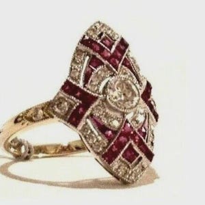 Può includere: Un anello d'oro ornato con un grande design geometrico. L'anello presenta una pietra centrale, rotonda e trasparente, circondata da pietre trasparenti più piccole e pietre rettangolari rosso scuro. La fascia è di un delicato colore oro.