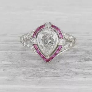 Könnte beinhalten: Ein Ring im Vintage-Stil mit einem birnenförmigen, klaren Edelstein in der Mitte, umgeben von einem rosa und weißen Design. Der Ring hat ein silbernes Band und ist mit kleinen, klaren Edelsteinen besetzt. Der Ring wird auf einer hellgrauen Oberfläche präsentiert.