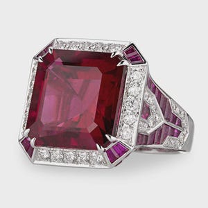 May include: A statement ring featuring a large, rectangular ruby gemstone. The ring has a white metal setting, possibly platinum or white gold, and is embellished with diamonds and smaller rubies. The band has a geometric design.
