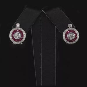 May include: A pair of round stud earrings with a central clear stone surrounded by a ring of red stones and a border of smaller clear stones. The earrings are displayed on a black stand against a black background.