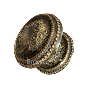 Può includere: Due pomelli per porte antichi in ottone con dettagli floreali ornati. I pomelli ovali presentano un motivo centrale in rilievo e un bordo perlato. L'ottone ha una calda patina invecchiata. La parola "Brass" è visibile nell'immagine.