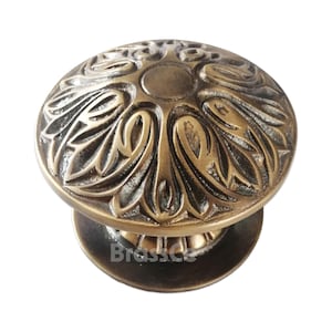 Puede incluir: Un pomo de armario de color bronce con un diseño floral ornamentado. El pomo tiene una parte superior redondeada con patrones detallados de hojas y pétalos, y una pequeña base circular. La palabra "Brassce" es visible en la base.