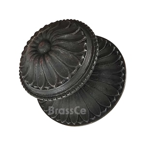 May include: Close-up of two antique-style, dark metal cabinet knobs. Each knob features a floral design with petal-like details and a central circular element. The knobs are likely made of cast iron or a similar material, with a weathered finish. The text "BrassCe" is visible.