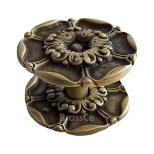 Könnte beinhalten: Antiker Türknauf aus Messing mit floralem Design. Der Knauf hat ein detailliertes, symmetrisches Muster mit einer zentralen Rosette und kunstvollen Schnörkeln. Der Sockel des Knaufs ist ebenfalls mit ähnlichen floralen Elementen verziert. Der Text "BrassCe" ist sichtbar.