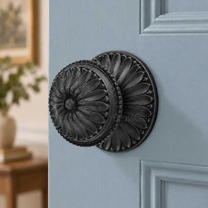 May include: A dark, matte black door knob featuring a circular, petal-like floral design on both the knob and the matching circular backplate. The hardware is mounted on a light blue paneled door, showcasing a textured, weathered finish.