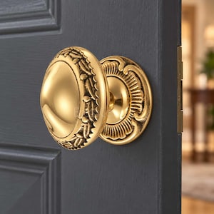 Può includere: Un pomello per porta in ottone dorato lucido con un centro liscio e arrotondato e un bordo decorativo testurizzato caratterizzato da un motivo a foglie ripetuto. Il pomello è montato su una rosetta circolare abbinata con bordo smerlato su una porta grigio scuro.