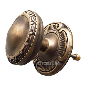 May include: Two antique brass door knobs with an aged finish. The knobs have an oval shape with intricate floral and leaf detailing around the edges. The backplate of one knob has the text "BrassCo".