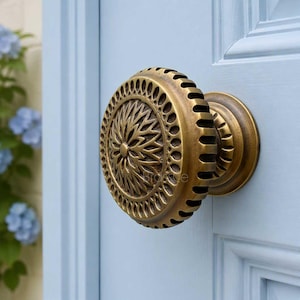 May include: A round brass door knob with an intricate, embossed floral pattern on the face and a ribbed edge. The knob is mounted on a light blue paneled door. The metal has a warm, aged finish with darker tones in the recessed areas.