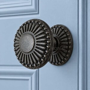 May include: A round, dark metallic door knob with a sunburst pattern radiating from a smooth center circle. The edge features a beaded border detail. The knob is mounted on a light blue panel with a rectangular frame design. The text BrassCe is visible.