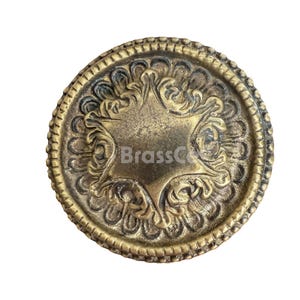 May include: A round, antique brass-colored decorative knob with a star-shaped center and ornate floral detailing. The knob has a textured, aged appearance and the text "BrassCo" is visible.