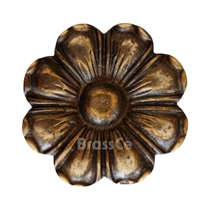 Könnte beinhalten: Ein detailliertes, bronzefarbenes, blütenförmiges Dekorationselement. Das Design weist einen zentralen, kreisförmigen Kern mit strahlenförmigen Blütenblättern auf. Die Oberfläche hat ein strukturiertes Aussehen, mit dem Text "BrassCe".