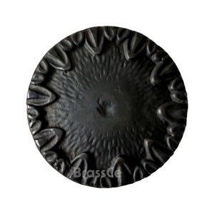 Può includere: Un bottone circolare in metallo scuro con una superficie strutturata e un bordo decorativo. Il bottone ha un'indentazione centrale e un motivo di forme stilizzate a forma di petalo attorno al perimetro. Il testo "BrassDe" è visibile in basso a sinistra.