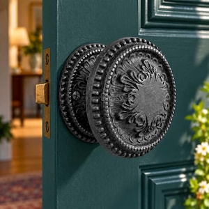 May include: A round, dark metallic door knob with an intricate, raised floral relief pattern and a beaded outer edge. The knob is mounted on a deep teal door, featuring a rectangular brass-coloured latch plate on the side edge of the door panel.