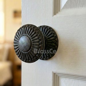 May include: A pair of antique-style, dark bronze door knobs. Each knob features a circular design with a central disc and radiating lines, mounted on a white door. The knobs have a weathered appearance, suggesting age and vintage charm.
