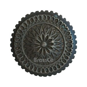 May include: A round, antique-style metal knob with a detailed floral design in the center and a decorative border. The knob is dark gray with a textured surface. The text "BrassCe" is visible on the knob.