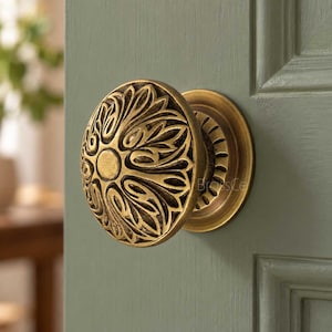May include: A round brass door knob featuring an intricate, embossed floral pattern with a central circular medallion. The metal has a warm, aged finish with darker recessed areas that highlight the detailed, symmetrical leaf-like carvings.