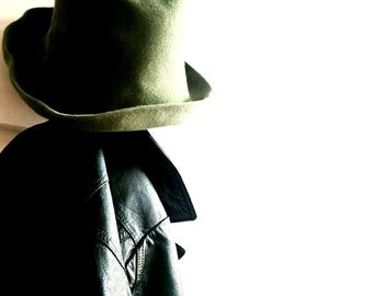 以下が含まれることがあります： オリーブグリーンのフェルトハットは、つばが広くわずかにカーブしており、黒いレザージャケットの上に配置されています。ジャケットには目に見える縫い目、構造化された襟、滑らかで反射する表面の質感があり、マットなフェルトハットと対照的です。