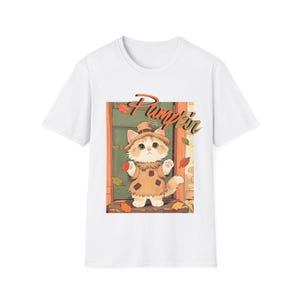 Puede incluir: Una camiseta blanca de manga corta con un estampado gráfico de un gatito esponjoso vestido de espantapájaros con un traje y sombrero color canela. El gatito está frente a una puerta con hojas de otoño. La palabra Pumpkin está escrita en naranja.