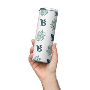 Puede incluir: Un vaso blanco con un patrón de hojas verdes y "VB". El vaso de acero inoxidable tiene tapa y está diseñado para mantener las bebidas frías o calientes. El diseño presenta un patrón repetido de hojas de monstera verdes y las letras "VB".