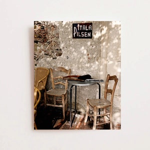 May include: A rustic cafe setting with wooden chairs and a small metal table. A sign reading "ITALA PILSEN" is mounted on a weathered stone wall. The scene is illuminated by sunlight, casting shadows.