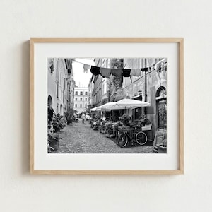May include: A black and white framed photograph depicting a narrow cobblestone street in Italy. The scene features laundry hanging on a line between buildings, outdoor cafe tables with white umbrellas, potted plants, and a bicycle parked nearby.