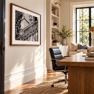 May include: A framed black and white photograph of the Notre-Dame cathedral exterior, featuring a rose window and stone statues, mounted on a light-colored wall in a room with a wooden desk, a black leather office chair, and a bookshelf.