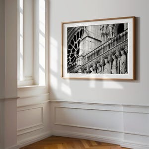 May include: A black and white framed wall art print depicting the architectural details of the Notre-Dame Cathedral in Paris, featuring a prominent rose window and a row of stone statues of figures standing along the gallery of the facade.