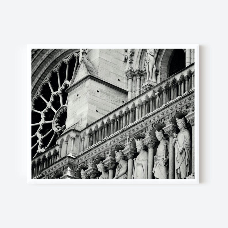 May include: A black and white architectural photograph of the Notre-Dame Cathedral facade, featuring a large circular rose window, stone columns, and a row of carved stone statues of figures standing in niches along the gallery of kings.