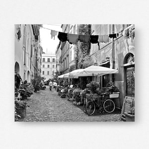 May include: A black and white street scene featuring a narrow cobblestone alleyway lined with tall buildings. Laundry hangs on a line stretched between the structures. A bicycle is parked near outdoor dining tables under white umbrellas.