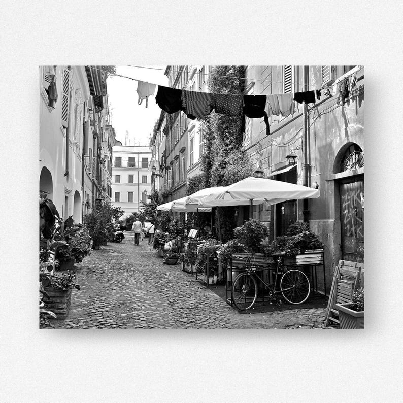 May include: A black and white street scene featuring a narrow cobblestone alleyway lined with tall buildings. Laundry hangs on a line stretched between the structures. A bicycle is parked near outdoor dining tables under white umbrellas.