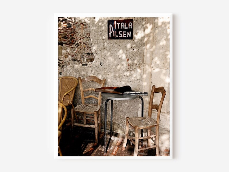 May include: An outdoor scene with a sign reading "ITALA PILSEN" on a weathered stone wall. A small metal table is surrounded by wooden chairs. Sunlight casts shadows across the scene, creating a rustic atmosphere.