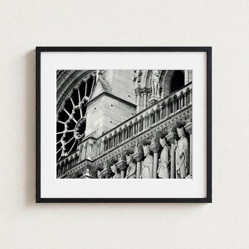A black and white framed photograph of the Notre-Dame Cathedral exterior, showcasing a detailed stone rose window, a series of arched columns, and a row of carved statues standing in niches along the building's facade.