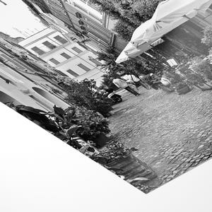 May include: A black and white photographic print showing a tilted perspective of a cobblestone street lined with buildings, outdoor umbrellas, and potted plants. The scene captures a narrow urban alleyway with architectural details and foliage.