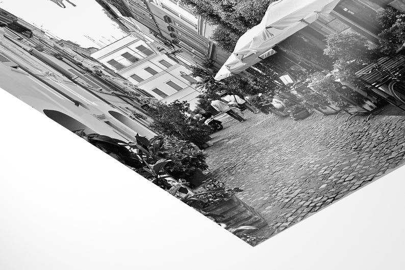 May include: A black and white photographic print showing a tilted perspective of a cobblestone street lined with buildings, outdoor umbrellas, and potted plants. The scene captures a narrow urban alleyway with architectural details and foliage.