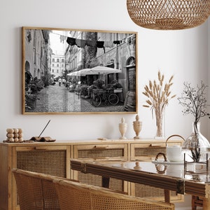 May include: A rectangular wall art print in a wooden frame featuring a black and white photograph of a narrow European cobblestone street with laundry hanging overhead, outdoor cafe umbrellas, potted plants, and a bicycle parked on the side.