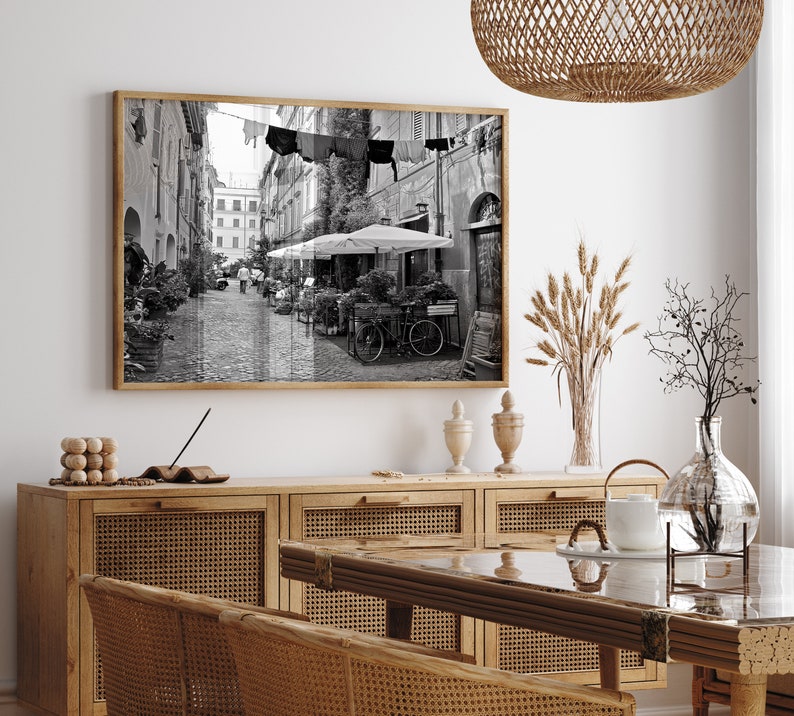 May include: A rectangular wall art print in a wooden frame featuring a black and white photograph of a narrow European cobblestone street with laundry hanging overhead, outdoor cafe umbrellas, potted plants, and a bicycle parked on the side.