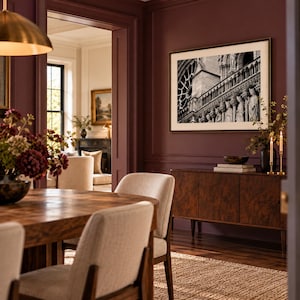 May include: A dining room featuring a wooden table with beige upholstered chairs, a dark wood sideboard, and a black and white framed photograph of Notre Dame cathedral hanging on a deep burgundy wall with decorative molding.