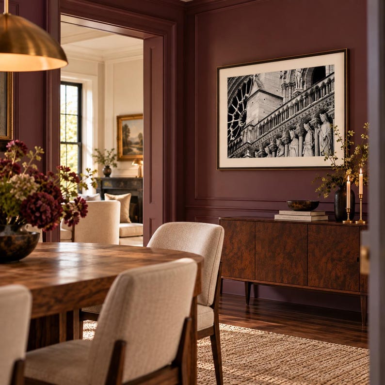 May include: A dining room featuring a wooden table with beige upholstered chairs, a dark wood sideboard, and a black and white framed photograph of Notre Dame cathedral hanging on a deep burgundy wall with decorative molding.
