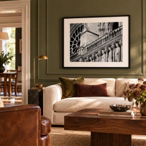 May include: A black and white framed photograph of the Notre Dame cathedral facade featuring a rose window and stone statues, displayed on an olive green wall above a beige sofa with velvet pillows and a wooden coffee table.