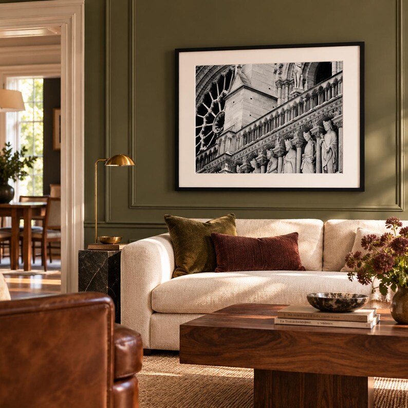 May include: A black and white framed photograph of the Notre Dame cathedral facade featuring a rose window and stone statues, displayed on an olive green wall above a beige sofa with velvet pillows and a wooden coffee table.