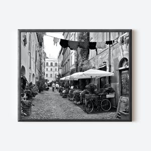 May include: A black and white photograph of a narrow cobblestone street lined with buildings. Laundry hangs on a line above the street. A bicycle is parked near white outdoor umbrellas and potted plants. People walk in the distance.
