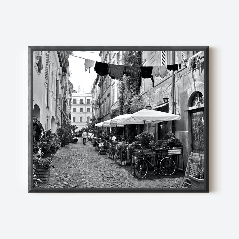 May include: A black and white photograph of a narrow cobblestone street lined with buildings. Laundry hangs on a line above the street. A bicycle is parked near white outdoor umbrellas and potted plants. People walk in the distance.