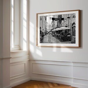 May include: A black and white framed wall art print depicting a narrow cobblestone street scene with laundry hanging on a line between buildings, outdoor cafe umbrellas, potted plants, and a bicycle parked along the side of the stone structures.